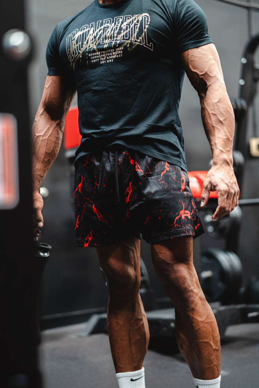 A muscular man in a gym wearing a black t-shirt with a graphic print and black athletic shorts featuring a red abstract pattern.
