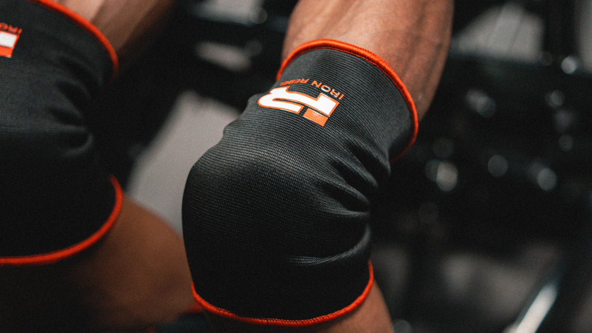 Close-up of a person lying on a gym bench pressing, wearing black knee sleeves with orange trim and a visible logo, along with a black shirt in a gym setting.