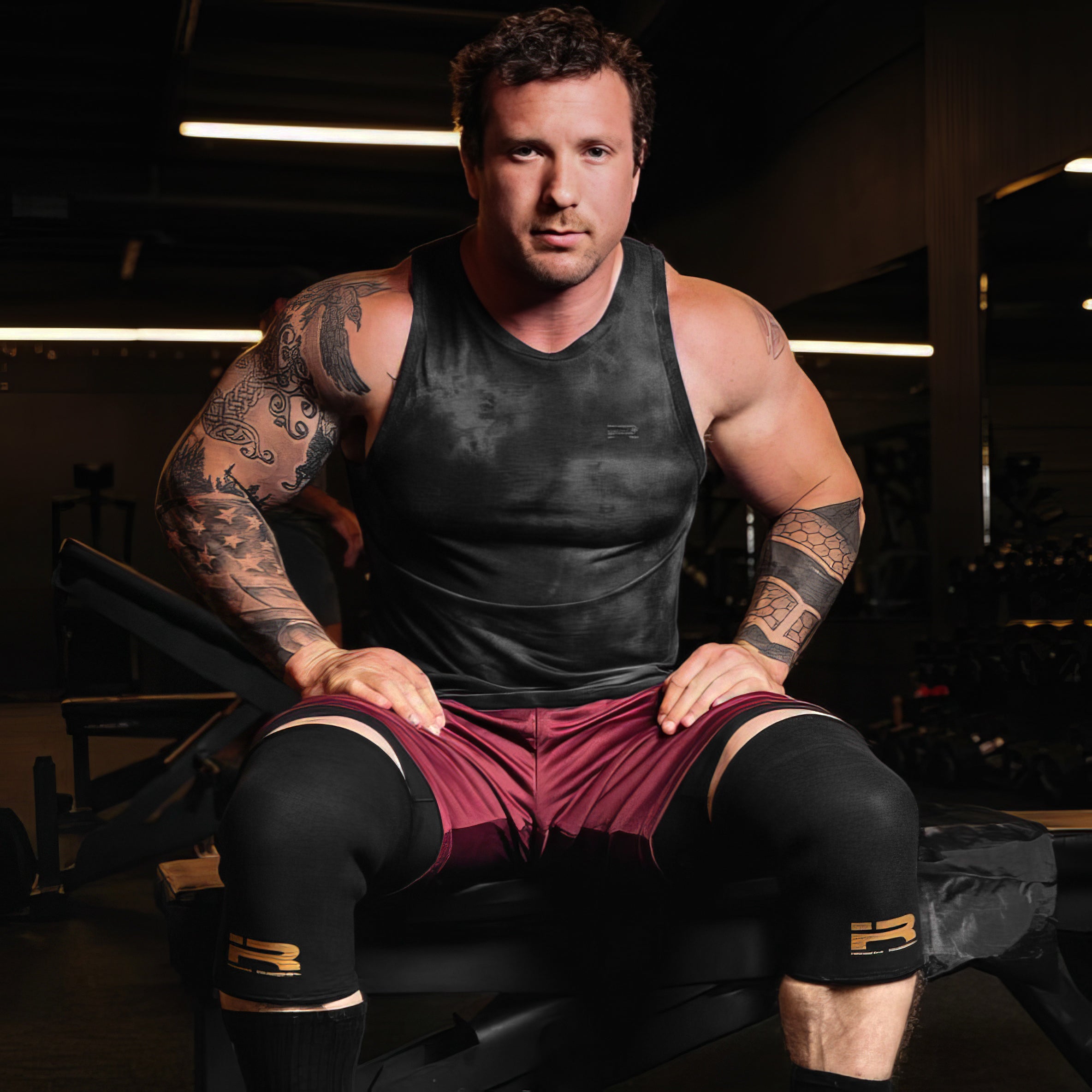Tattooed male lifter wearing black Iron Rebel PR Cone Knee Sleeves with a gold logo while sitting on a bench in the gym. Shows the fit and compression of the sleeves during heavy weightlifting and powerlifting workouts.